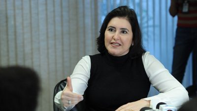 Simone Tebet dá entrevista sentada, com vários microfones na mesa em sua frente. Eal veste camisa branca e blusa sem mangas preta.
