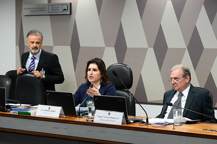 Homem de pé veste terno preto e segura em cadeira preta. No centro, Simone Tebet está sentada com notebook a frente. Do outro lado, homem veste terno cinza escuro. Ele também está sentado