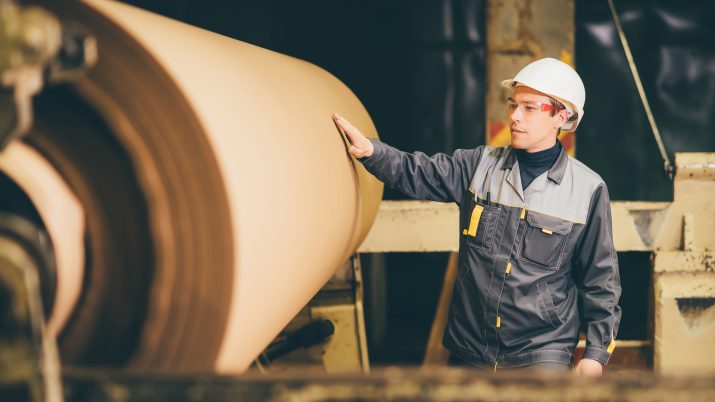 Homem usando equipamentos de proteção individual com a mão direita numa bobina de papel; imagem simboliza as empresas do setor, como Suzano (SUZB3) e Klabin (KLBN11)