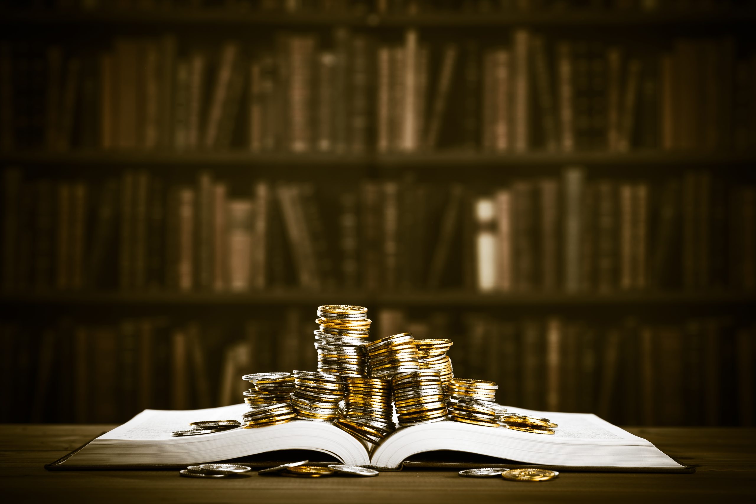 Moedas empilhadas em cima de um livro aberto. Ao fundo, uma estante.