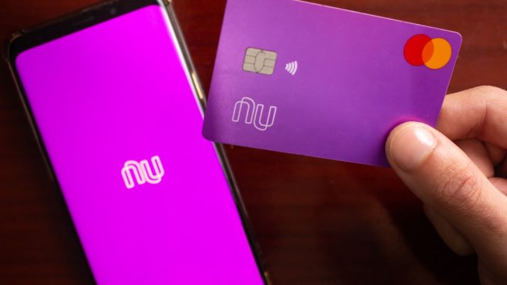 Homem segurando um smartphone com o aplicativo do banco na tela e o cartão Nubank (NU/NUBR33) na mesa