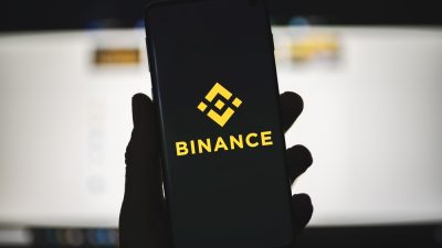 Uma mão segura um smartphone com a tela preta e o logo da corretora Binance em amarelo no centro
