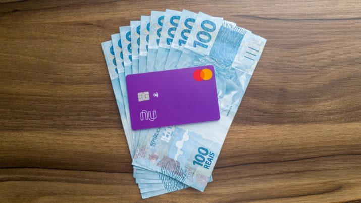 Cartão do Nubank sobre várias notas de 100 reais em cima de uma mesa de madeira