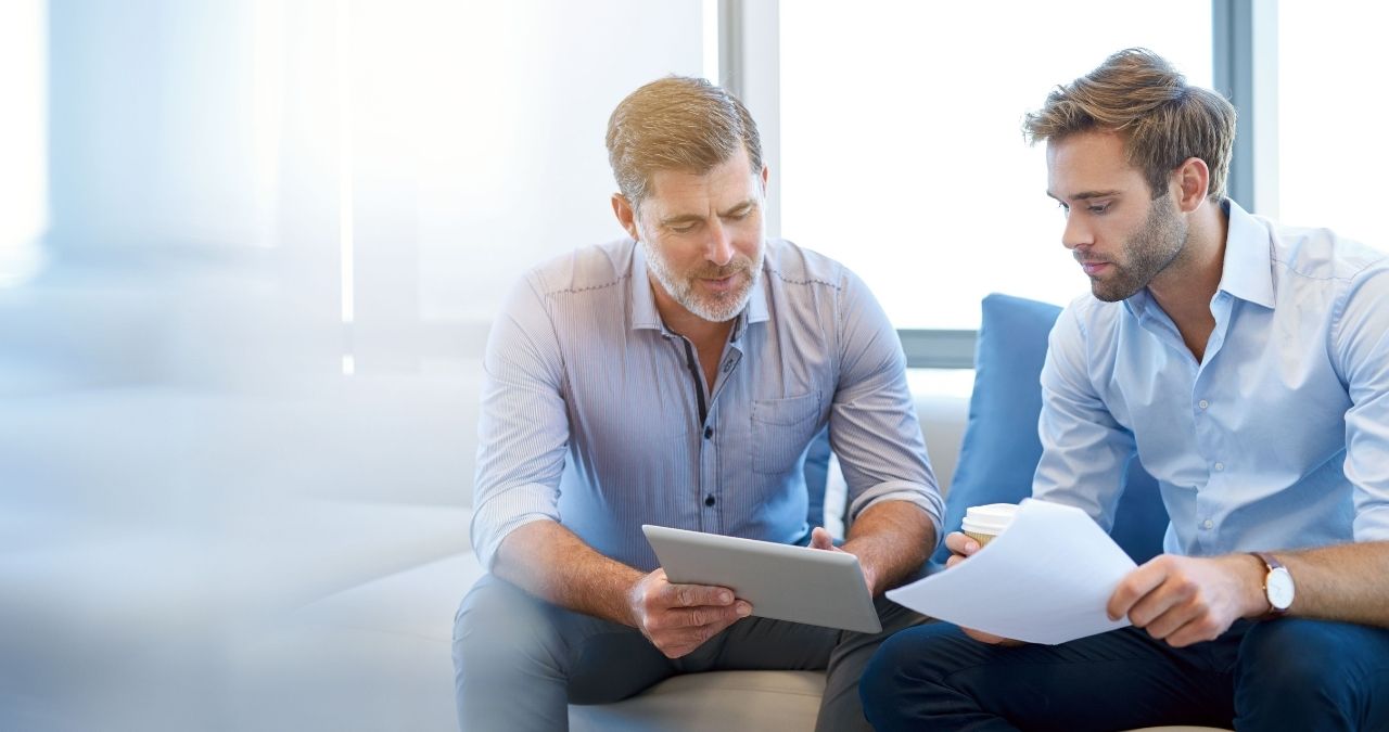 Dois homens brancos de terno conversando; um com um tablet na mão, outro com papéis na mão, representando soft skills