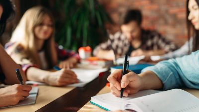 Mão escrevendo em um caderno com alunos desfocados ao fundo