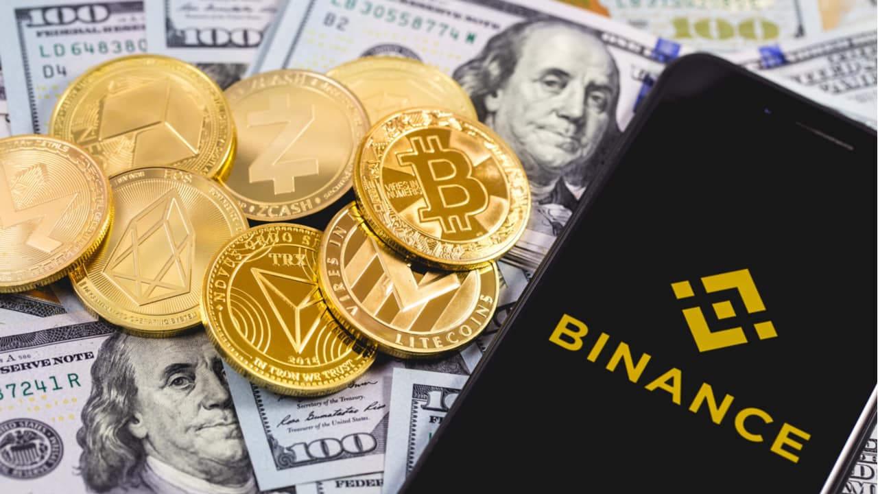 Foto de criptomoedas jogadas em cima de notas de dólar, ao lado de um celular mostrando o logo da Binance