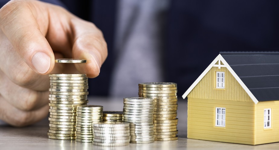 Moedas empilhadas ao lado de uma pequena casa amarela representando o investimento em fundos imobiliários | Maxi Renda HCTR11 fii fiis