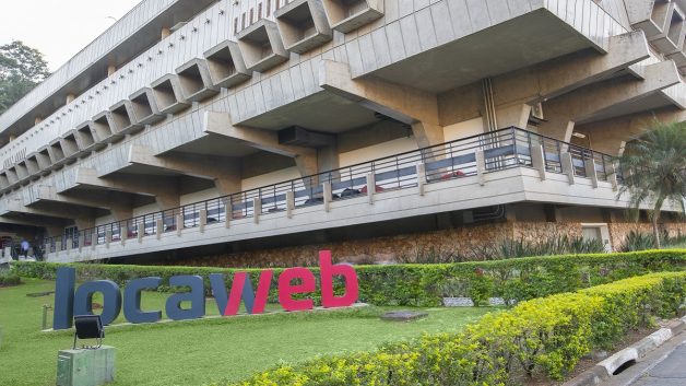 Fachada de um prédio com um letreiro da Locaweb (LWSA3) no primeiro plano, em cima de um gramado