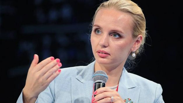 Mulher loira, de cabelos presos, segura microfone enquanto fala gesticulando com a outra mão