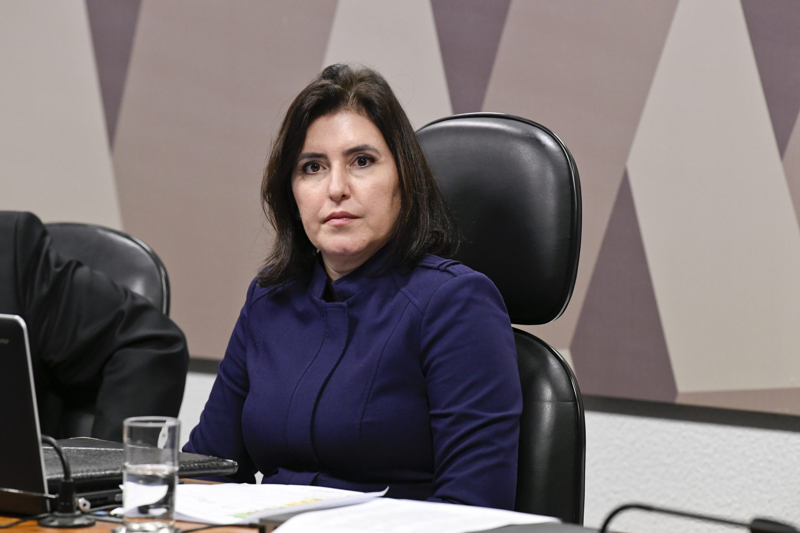 Simone Tebet veste blusa azul marinho. Ela está sentada em uma cadeira preta, com um notebook em sua frente e um copo de água ao lado.