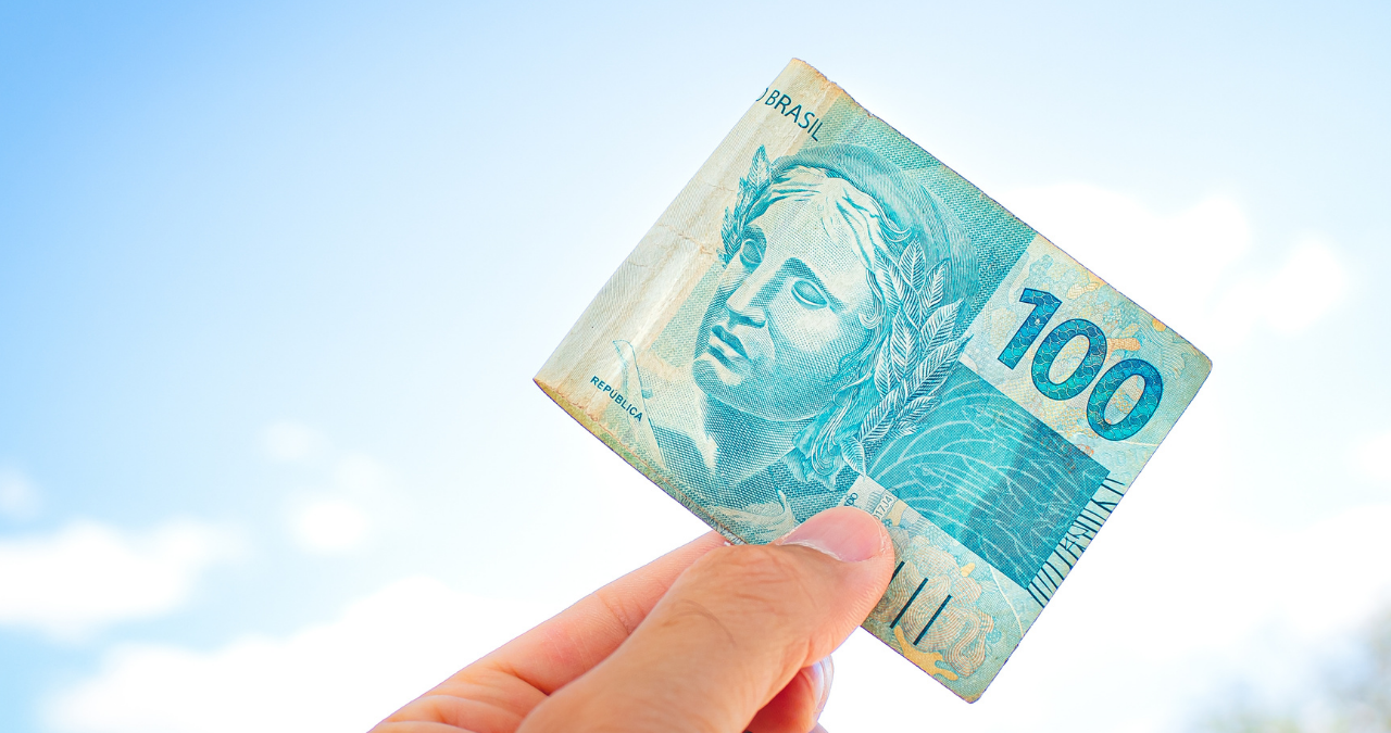 Mão segura nota de 100 reais. representando "bônus" do Governo. Dívidas
