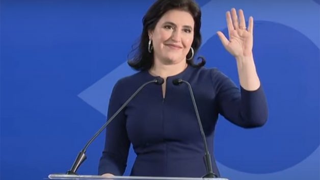 Simone Tebet veste roupa azul escura. De pé, ela está em um púlpito e acena com uma das mãos.