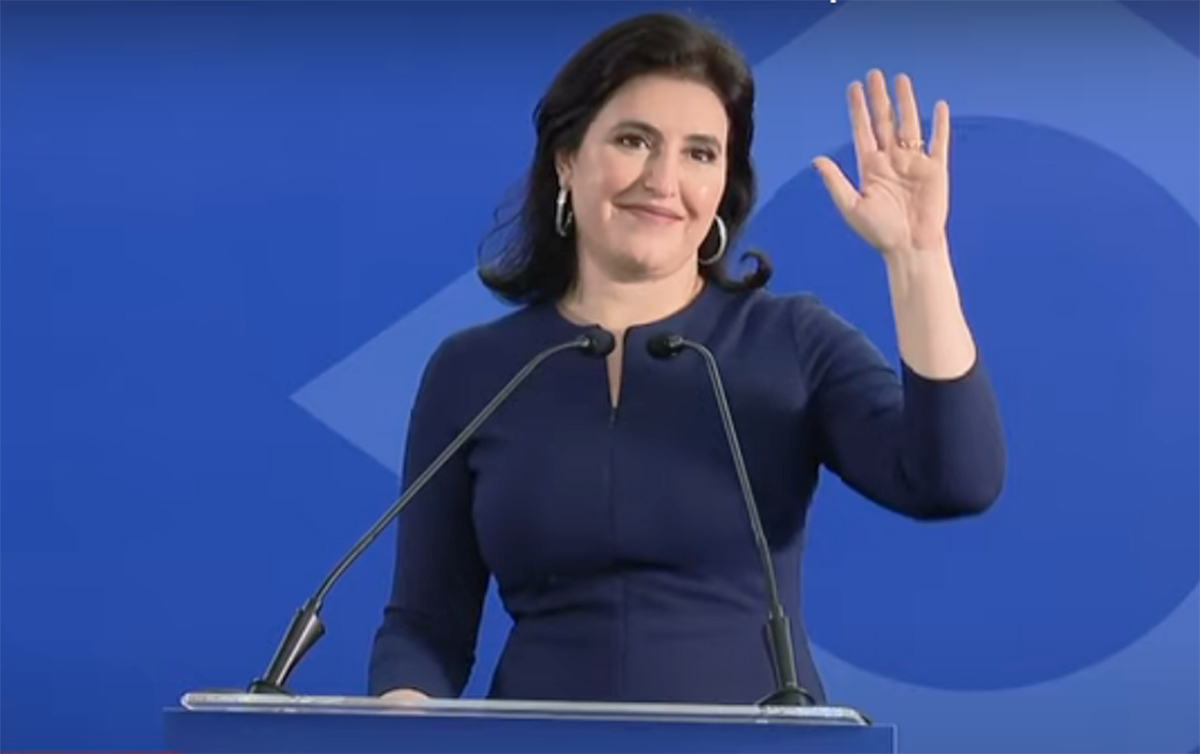Simone Tebet veste roupa azul escura. De pé, ela está em um púlpito e acena com uma das mãos.