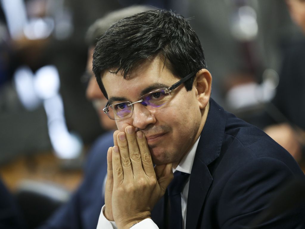 O senador Randolfe Rodrigues com as mãos juntas e próximas à boca, apoiado em uma bancada