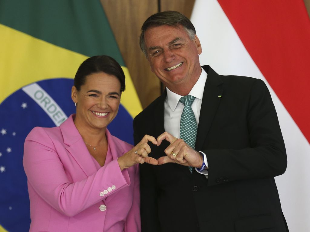 A presidente da Hungria veste rosa e faz um coração com uma das mãos junto com o presidente Jair Bolsonaro. Bandeiras de Hungria e Brasil ao fundo