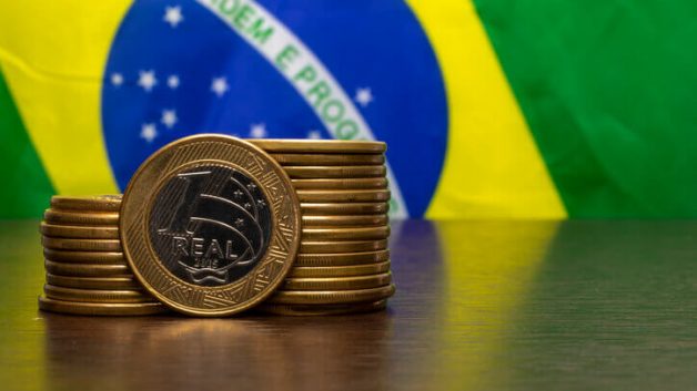 Moedas de real com bandeira do Brasil ao fundo