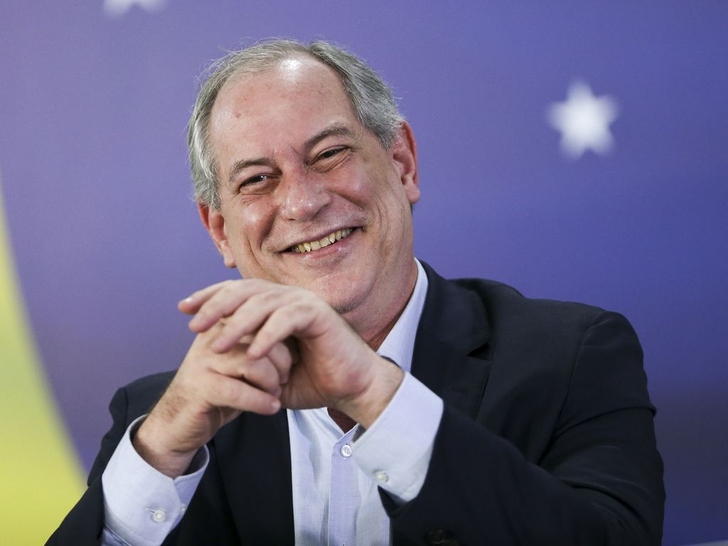 Foto de meio corpo de Ciro Gomes sorrindo com a bandeira do Brasil ao fundo
