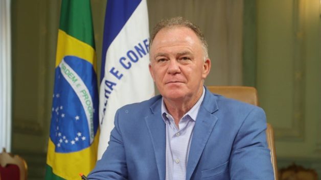 Homem vestido de blazer azul e camisa azula mais clara com bandeira do Brasil e do ES atrás e um óculos de grau sobre a mesa