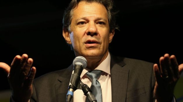 Fernando Haddad fala em microfone, com os braços abertos. Ele veste terno cinza, camisa clara e gravata azul.