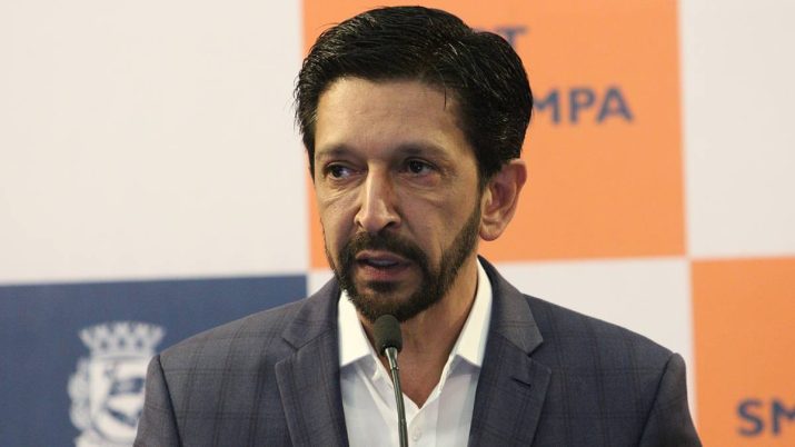 O prefeito de São Paulo, Ricardo Nunves, fala ao microfone com um painel azul, branco e laranja ao fundo em ato por moradia em são paulo