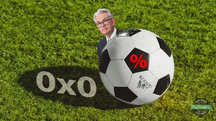 Em um gramado, aparece um placar com zero a xero, uma bola de futebol com uma % no centro e o presidente do Federal Reserve, Jerome Powell, escondido atrás da bola