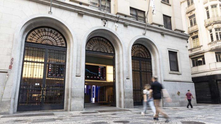 Fachada do prédio da B3. No interior, há painel com cotações. Duas pessoas andam em direção ao prédio.