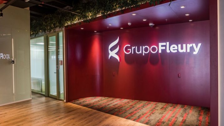 Parede vermelha vista da lateral. Há o logo e o nome do Grupo Fleury em led branco. Na lateral esquerda há portas de vidro e uma parede branca. Acima, há plantas.