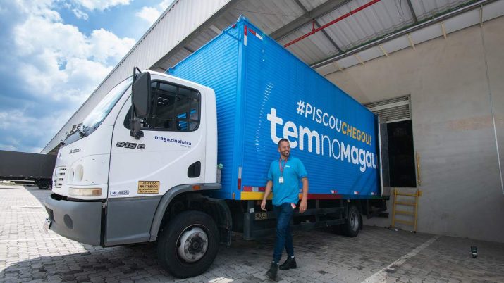 Centro de distribuição da Magazine Luiza em Guarulhos. Há um caminhão azul, com os dizeres "#PISCOUCHEGOU!" em branco e amarelo, e "tem no magalu" em branco. Na frente, há um funcionário com camisa da empresa.