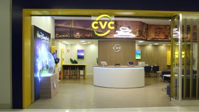 Agência da CVC com fachada azul e amarela, logo da companhia na entrada. Dentro, há cadeiras e balcões