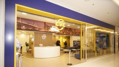 Agência da CVC com fachada azul e amarela, logo da companhia na entrada. Dentro, há cadeiras, balcões e pessoas conversando