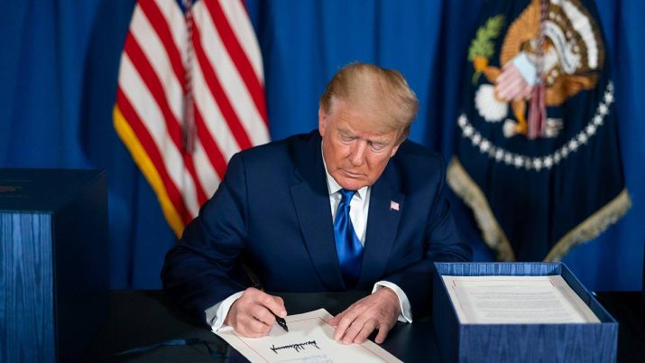 Donald Trump está sentado em frente a uma mesa, com uma caixa azul na lateral.