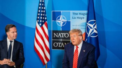 Imagem mostra Donald Trump vestindo terno azul e gravata vermelha. Ele está sentando em uma cadeira ao lado do presidente da Otan, Mark Rutte, que também usa terno escuro e camisa branca. De fundo, a bandeira dos EUA e o símbolo da Otan.