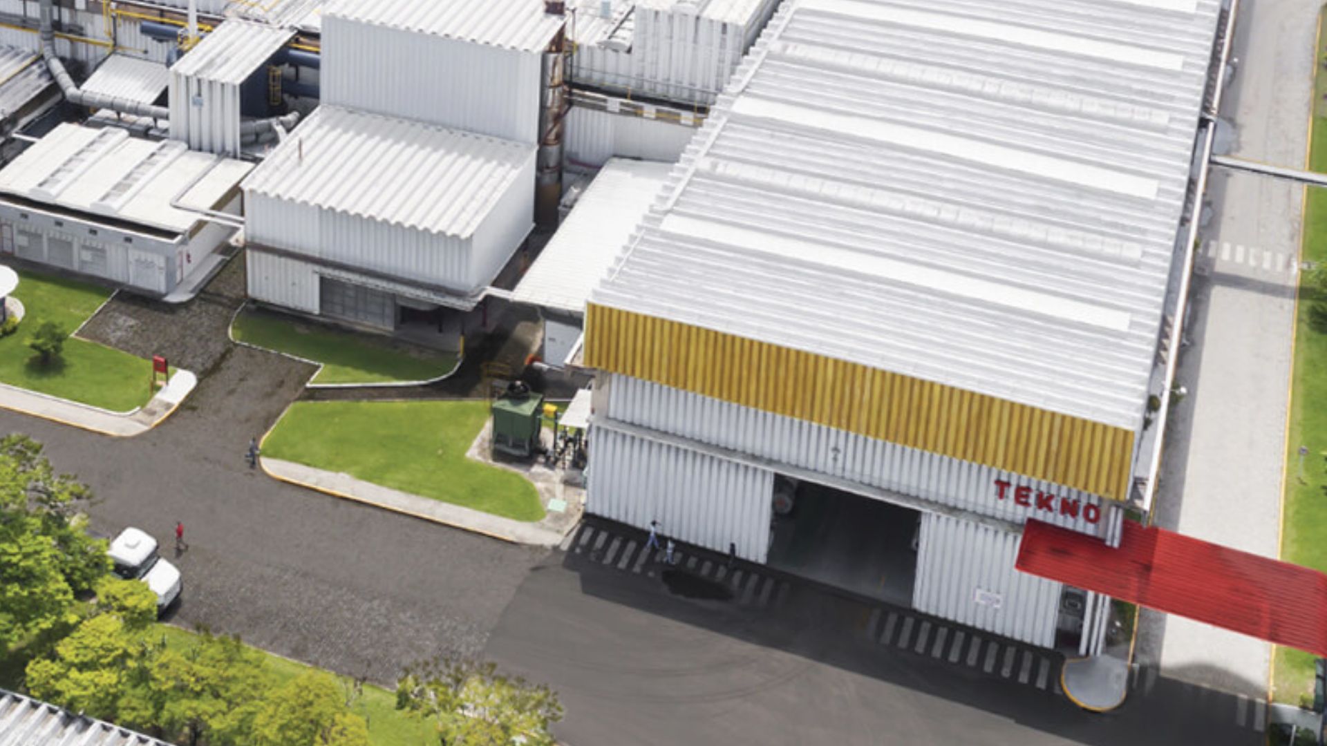 Vista aérea da fábrica da Tekno, com galpões industriais de telhado branco e detalhes em amarelo, cercada por áreas verdes e vias asfaltadas; na fachada, aparece a logomarca vermelha da empresa.