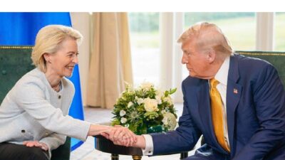 Foto mostra Ursula Von de Leyen sentada em uma cadeira, com a bandeira da UE ao fundo. Ela aperta as mãos de Donald Trump, que está sentado em uma cadeira ao seu lado e tem a bandeira dos EUA ao fundo.