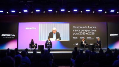 Evento Warren Day 2025 em auditório lotado, com painel de gestores de fundos discutindo perspectivas para 2025 e 2026. No palco, tdois participantes sentados em poltronas pretas: Bruno Cordeiro, Sócio da Kapitalo Investimentos e Felipe Guerra, da Legacy Capital. No telão ao fundo, Rogério Xavier participa da conversa via Zoom.