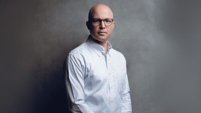 Retrato profissional de Roberto Valério, CEO da Cogna Educação. Ele é um homem careca de óculos, vestindo uma camisa xadrez clara. A imagem em estúdio tem um fundo cinza com um efeito esfumaçado e iluminação dramática, com o executivo olhando diretamente para a câmera com uma expressão séria