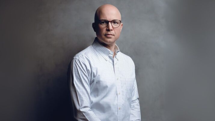 Retrato profissional de Roberto Valério, CEO da Cogna Educação. Ele é um homem careca de óculos, vestindo uma camisa xadrez clara. A imagem em estúdio tem um fundo cinza com um efeito esfumaçado e iluminação dramática, com o executivo olhando diretamente para a câmera com uma expressão séria