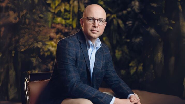 Roberto Valério, o CEO da Cogna Educação, posa para um retrato profissional. Na imagem, ele aparece sentado, de frente para a câmera, usando um blazer xadrez azul-marinho sobre uma camisa social azul clara. Óculos de aro fino e um corte de cabelo raspado definem sua aparência. O plano de fundo é sutilmente iluminado e desfocado, com a textura de folhagens e plantas em tons escuros e terrosos, sugerindo um ambiente sofisticado e natural. A fotografia é nítida, com foco em Valério, e destaca sua postura confiante e o olhar direto para o espectador.