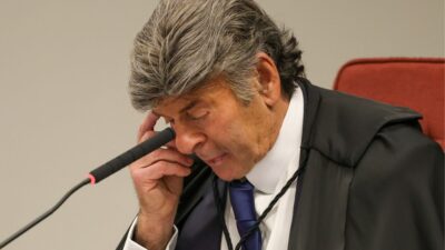 Luiz Fux, Ministro do Supremo Tribunal Federal (STF), durante o julgamento de Jair Bolsonaro e réus, mostrando um momento de reflexão enquanto se passa o julgamento. Fux aparece com a mão direita no rosto, ajustando seus óculos e visivelmente atento ao processo. Ele está usando um terno preto com a capa da toga, e ao fundo é possível ver a bancada do tribunal.