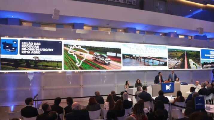Um grupo de pessoas sentadas em uma plateia observa o palco principal de um evento. O palco é dominado por um telão horizontal extralongo que exibe imagens de rodovias e paisagens rurais. No telão, estão em destaque as palavras "LEILÃO DAS RODOVIAS BR-060/364/GO/MT" e "ROTA AGRO", indicando um leilão de concessão de rodovias. No centro do palco, dois homens estão falando em um púlpito com o logotipo da B3 (bolsa de valores do Brasil). Uma mulher sentada à esquerda, na primeira fila, também está visível. O ambiente é formal, com iluminação azul e branca, e cadeiras brancas dispostas em fileiras para a audiência. Algumas pessoas na plateia estão usando crachás.