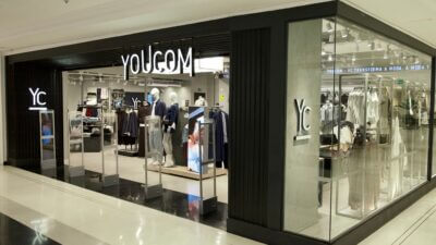 Loja da YouCom, destacando a fachada moderna com a marca 'YC' e o slogan 'YOUCOM - YC TRANSFORMA A MODA, A MODA TE TRANSFORMA'. O interior da loja é visível, com manequins e roupas expostas nas prateleiras