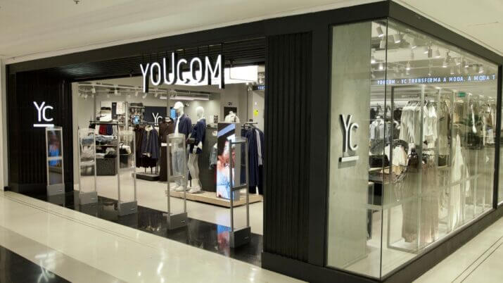 Loja da YouCom, destacando a fachada moderna com a marca 'YC' e o slogan 'YOUCOM - YC TRANSFORMA A MODA, A MODA TE TRANSFORMA'. O interior da loja é visível, com manequins e roupas expostas nas prateleiras