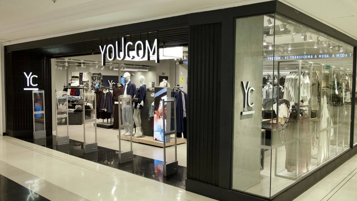 Loja da YouCom, destacando a fachada moderna com a marca 'YC' e o slogan 'YOUCOM - YC TRANSFORMA A MODA, A MODA TE TRANSFORMA'. O interior da loja é visível, com manequins e roupas expostas nas prateleiras