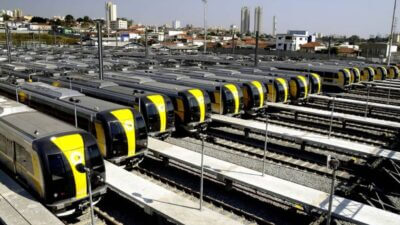 Vista aérea de um pátio de manutenção e estacionamento de trens da Linha 4-Amarela do Metrô de São Paulo. Dezenas de composições modernas, de cor cinza com faixas amarelas na frente e nas laterais, estão estacionadas lado a lado em várias linhas férreas. Os trens têm janelas frontais amplas e um design aerodinâmico. Ao fundo, pode-se ver a paisagem urbana com edifícios residenciais e comerciais de diferentes alturas, alguns mais antigos e outros mais modernos, sob um céu claro e ensolarado. A imagem transmite uma sensação de organização e infraestrutura de transporte público.