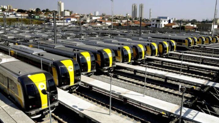 Vista aérea de um pátio de manutenção e estacionamento de trens da Linha 4-Amarela do Metrô de São Paulo. Dezenas de composições modernas, de cor cinza com faixas amarelas na frente e nas laterais, estão estacionadas lado a lado em várias linhas férreas. Os trens têm janelas frontais amplas e um design aerodinâmico. Ao fundo, pode-se ver a paisagem urbana com edifícios residenciais e comerciais de diferentes alturas, alguns mais antigos e outros mais modernos, sob um céu claro e ensolarado. A imagem transmite uma sensação de organização e infraestrutura de transporte público.