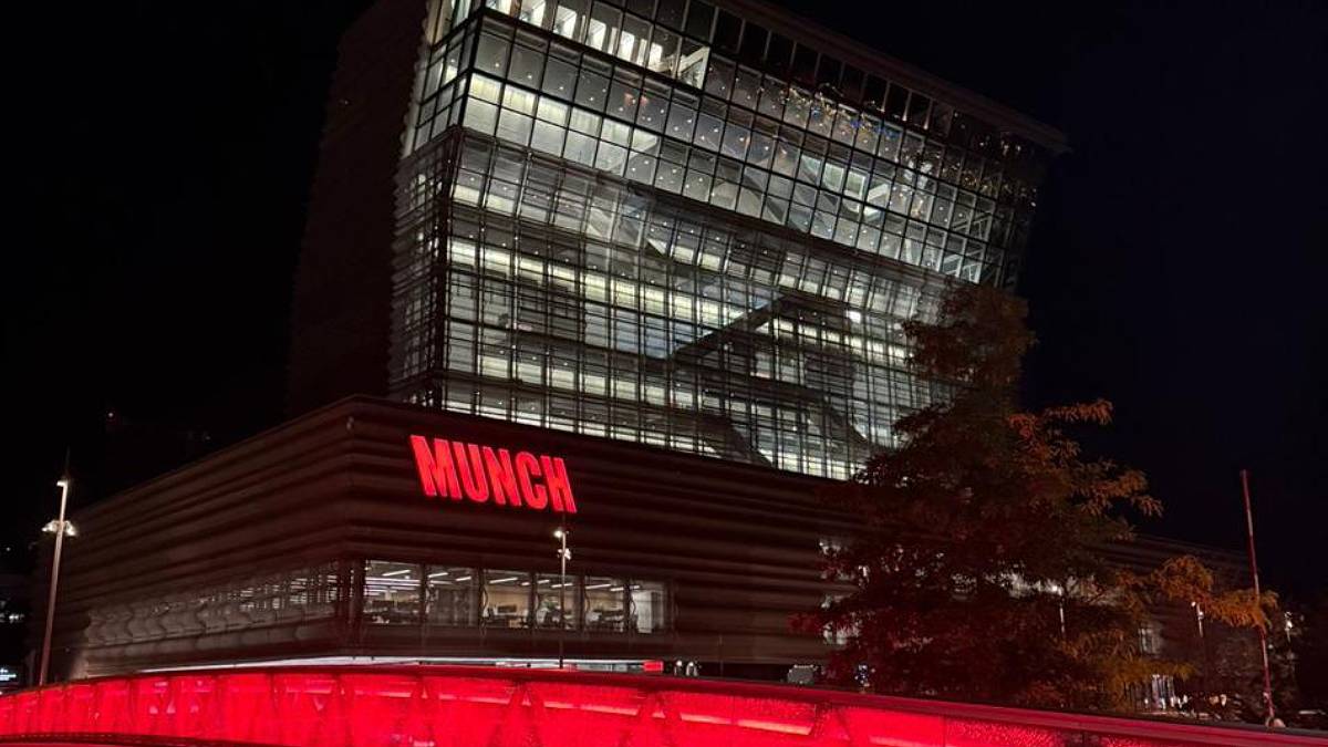 Foto noturna da fachada do Museu Munch, em Oslo, na Noruega. O prédio espelhado do museu é iluminado por uma luz vermelha, que destaca o nome do museu e a ponte em frente.
