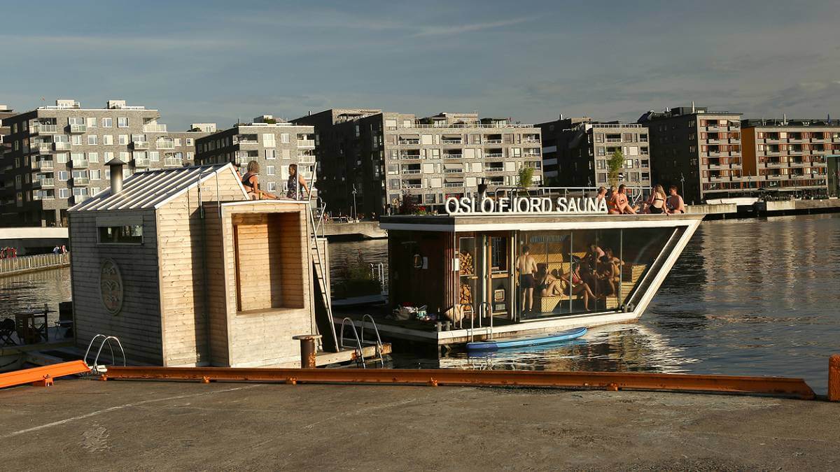 Imagem mostra um das saunas urbanas em Oslo, na Noruega. Na foto, um deck de madeira em primeiro plano, com a sauda flutuante e edifícios ao fundo.