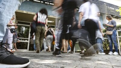 Candidatos chegam e se movimentam em frente a um prédio escolar antes da abertura dos portões para a aplicação das provas do CONCURSO PÚBLICO NACIONAL UNIFICADO (CNU 2025). Pessoas aparecem em pé e caminhando, algumas com mochilas e materiais na mão, em clima de expectativa no dia do exame