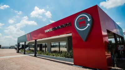 Fachada da unidade Vamos Seminovos, com estrutura moderna em vermelho e cinza, dedicada à venda de caminhões e máquinas usadas do Grupo Vamos, sob céu azul e dia ensolarado.
