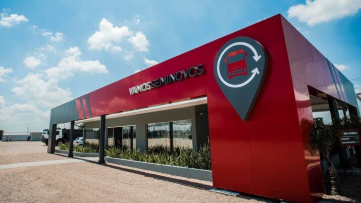 Fachada da unidade Vamos Seminovos, com estrutura moderna em vermelho e cinza, dedicada à venda de caminhões e máquinas usadas do Grupo Vamos, sob céu azul e dia ensolarado.